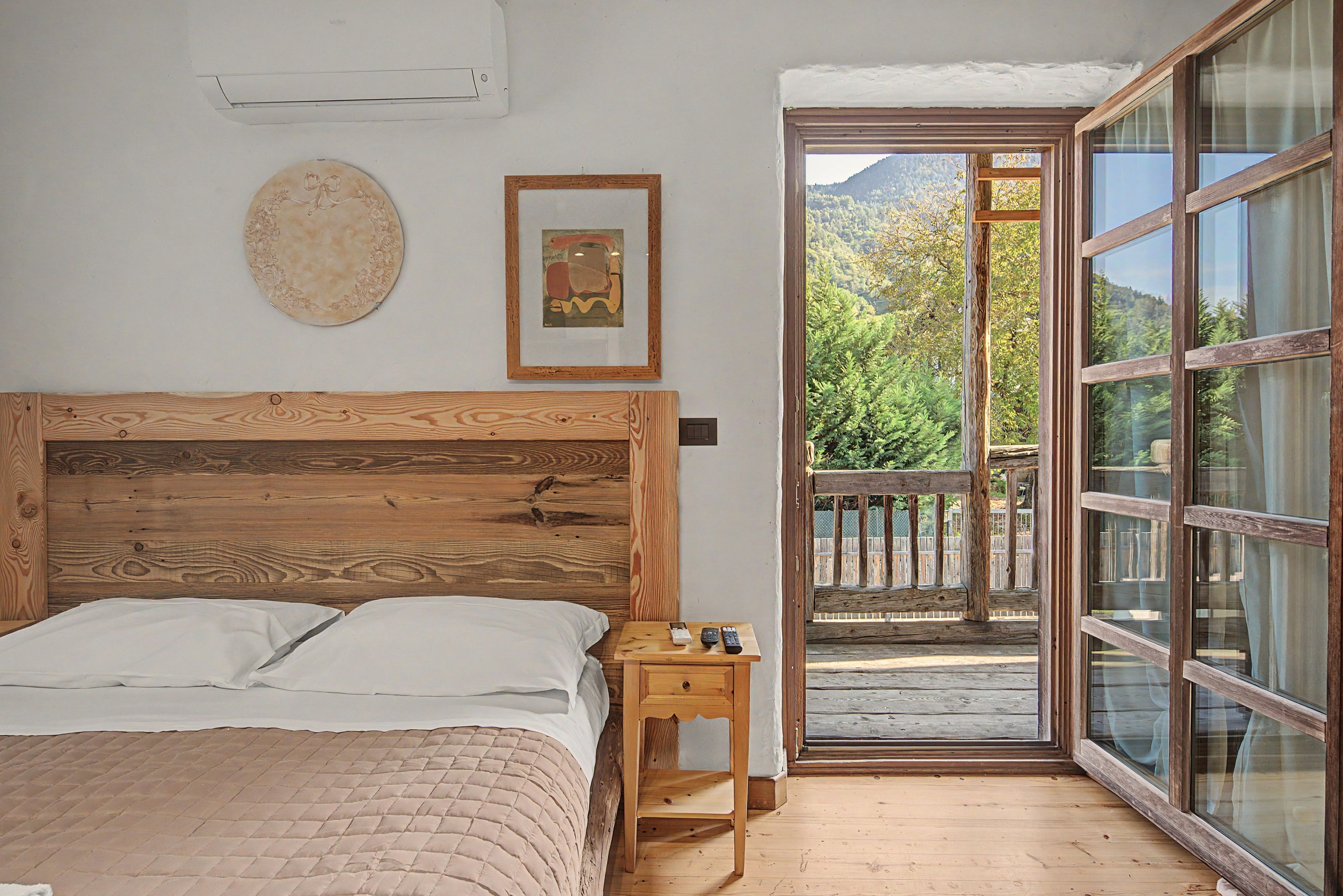 A bedroom with a wooden headboard, a bed with white linens, a small bedside table with a remote control, and a large window with a view of trees and mountains.