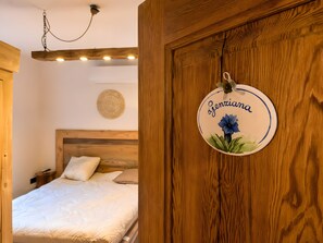 Un dormitorio con cabecera de madera, cama con ropa blanca, lámpara colgante y un letrero redondo sujeto a la pared con una flor y texto.