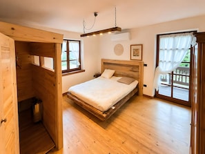 Habitació amb un llit de fusta, una finestra amb cortines, una làmpada al sostre i un petit safataire.