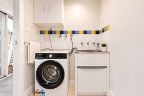 A compact laundry area with a front-loading washing machine, a sink, and a cabinet. - Coastal Cottage Getaway | Blue Bay, Central Coast (Blue Bay)