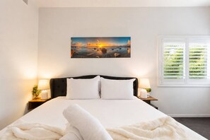 A bedroom with a bed, two bedside tables, a nightstand, a window with blinds, and a painting on the wall. - Coastal Cottage Getaway | Blue Bay, Central Coast (Blue Bay)