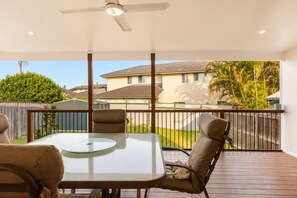 A covered patio with a table and chairs, a ceiling fan, and a view of a residential area with houses and greenery. - Coastal Cottage Getaway | Blue Bay, Central Coast (Blue Bay)