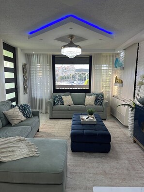 A modern living room with a grey sofa, a blue ottoman, and a view of a cityscape through a large window. - Your dream marina-front getaway is waiting for you (Fajardo city)