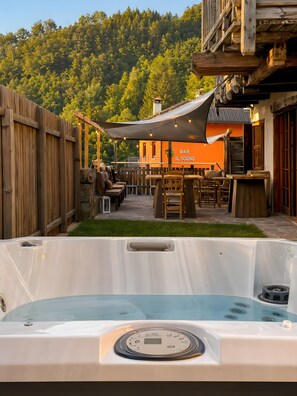 A hot tub in a backyard with a wooden fence, outdoor seating, and a building with an orange facade in the background.
