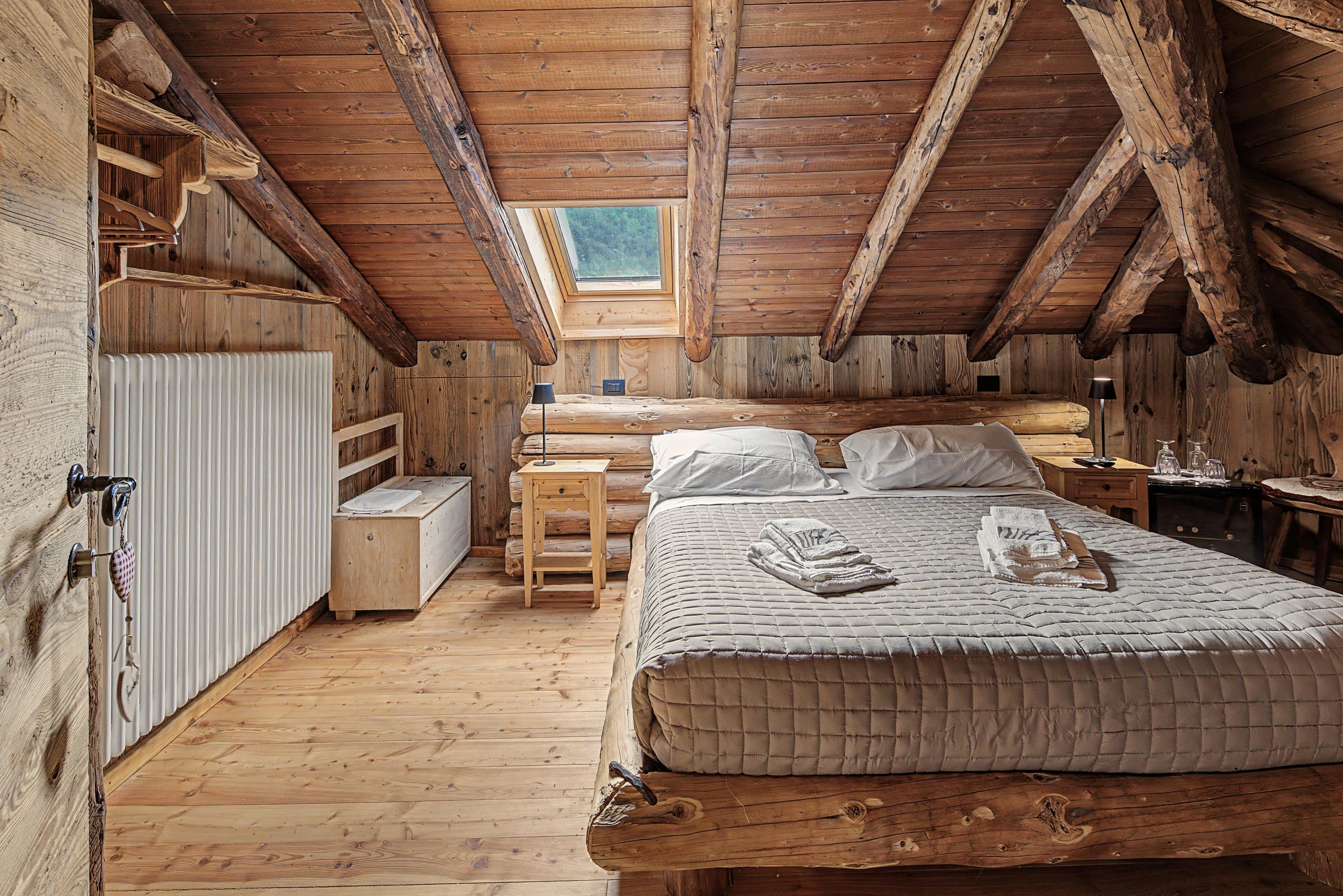Une chambre confortable située sous les toits, avec un plafond en pente, des murs en bois, un lit avec des oreillers, une table de chevet et une commode, ainsi qu’une fenêtre.