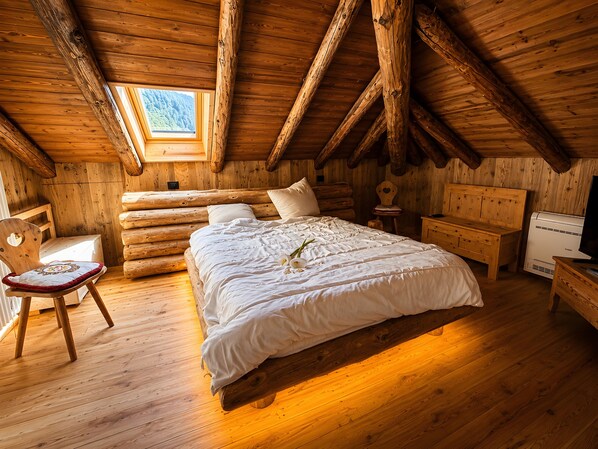 A log cabin bedroom with a wooden bed, a chair, a bedside table, and a window.
