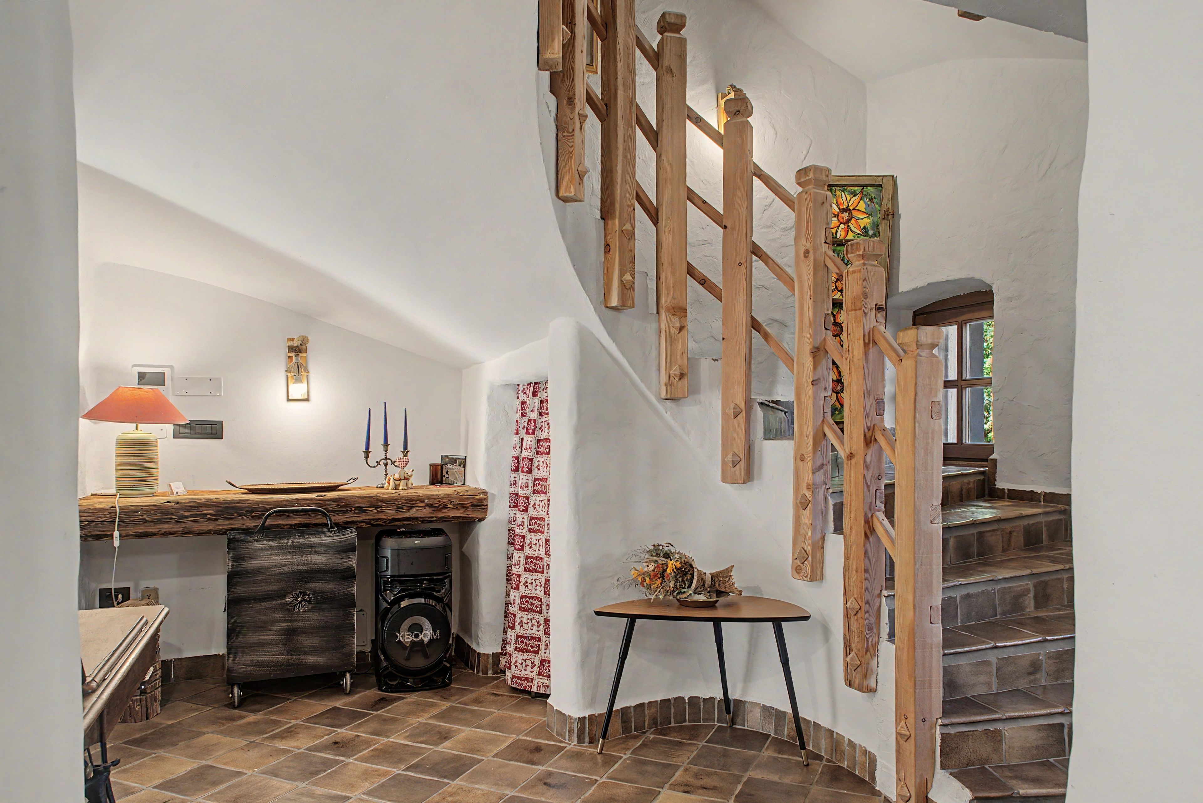 A rustic interior with a wooden staircase, a desk with a lamp, and a small table with a vase.
