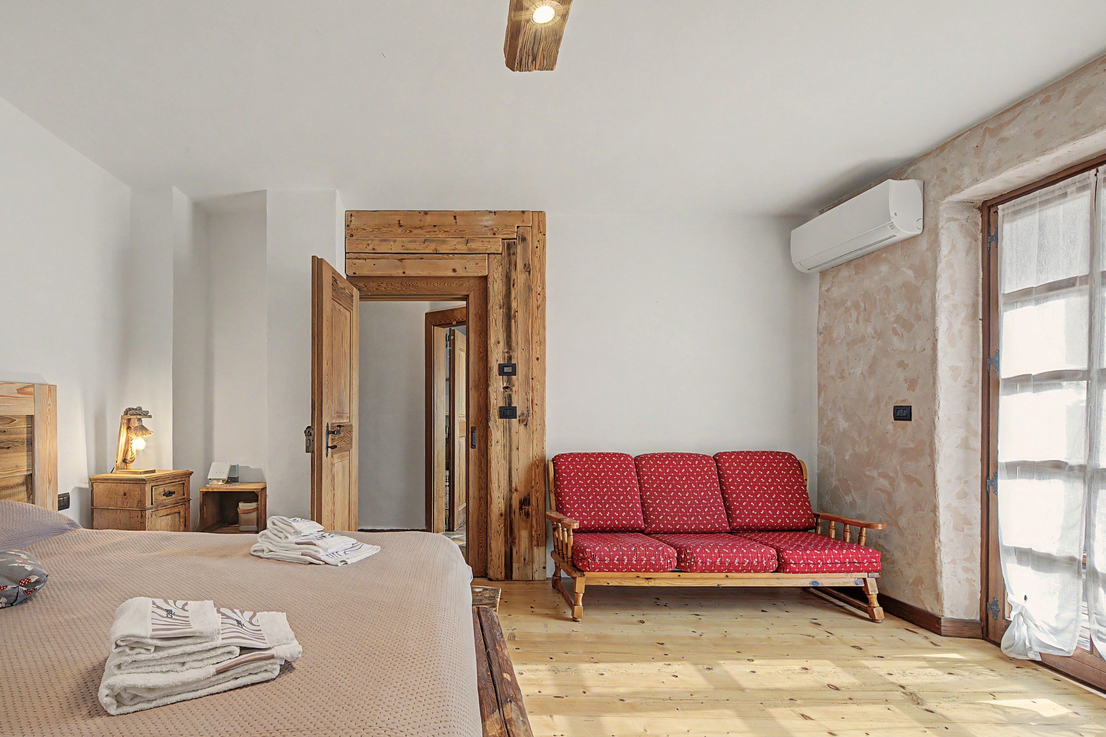 Ein Schlafzimmer mit einem Bett, einer Holzbank, einem roten, gemusterten Sofa und einer Holztür.