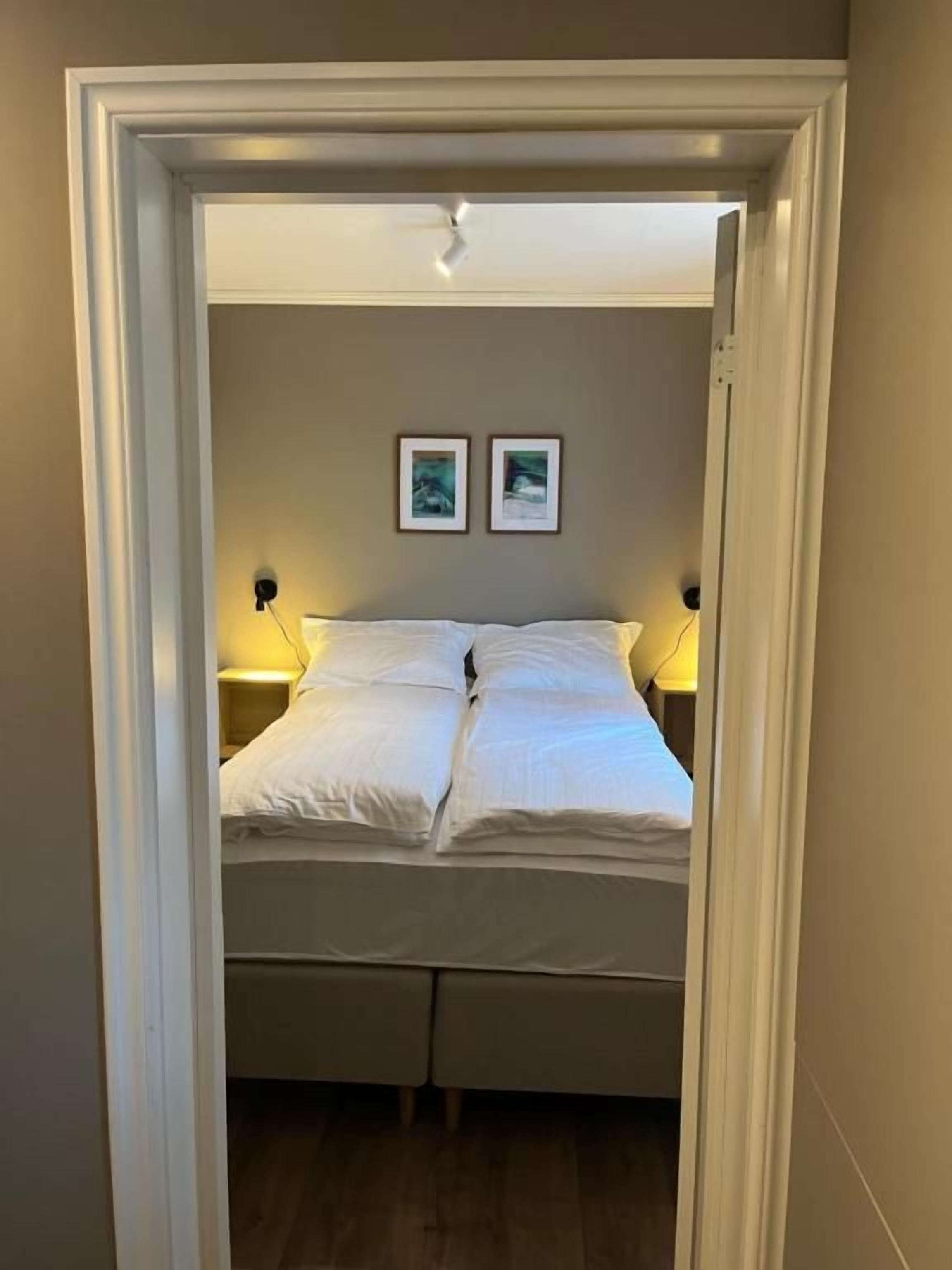A narrow doorway leading to a bed with white bedding, two wall-mounted nightstands, and two framed pictures on the wall.