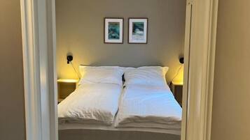 A narrow doorway leading to a bed with white bedding, two wall-mounted nightstands, and two framed pictures on the wall.