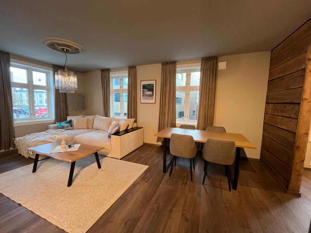A living room with a sofa, coffee table, dining table, and chairs. There are windows with curtains, a chandelier, and a wooden wall.