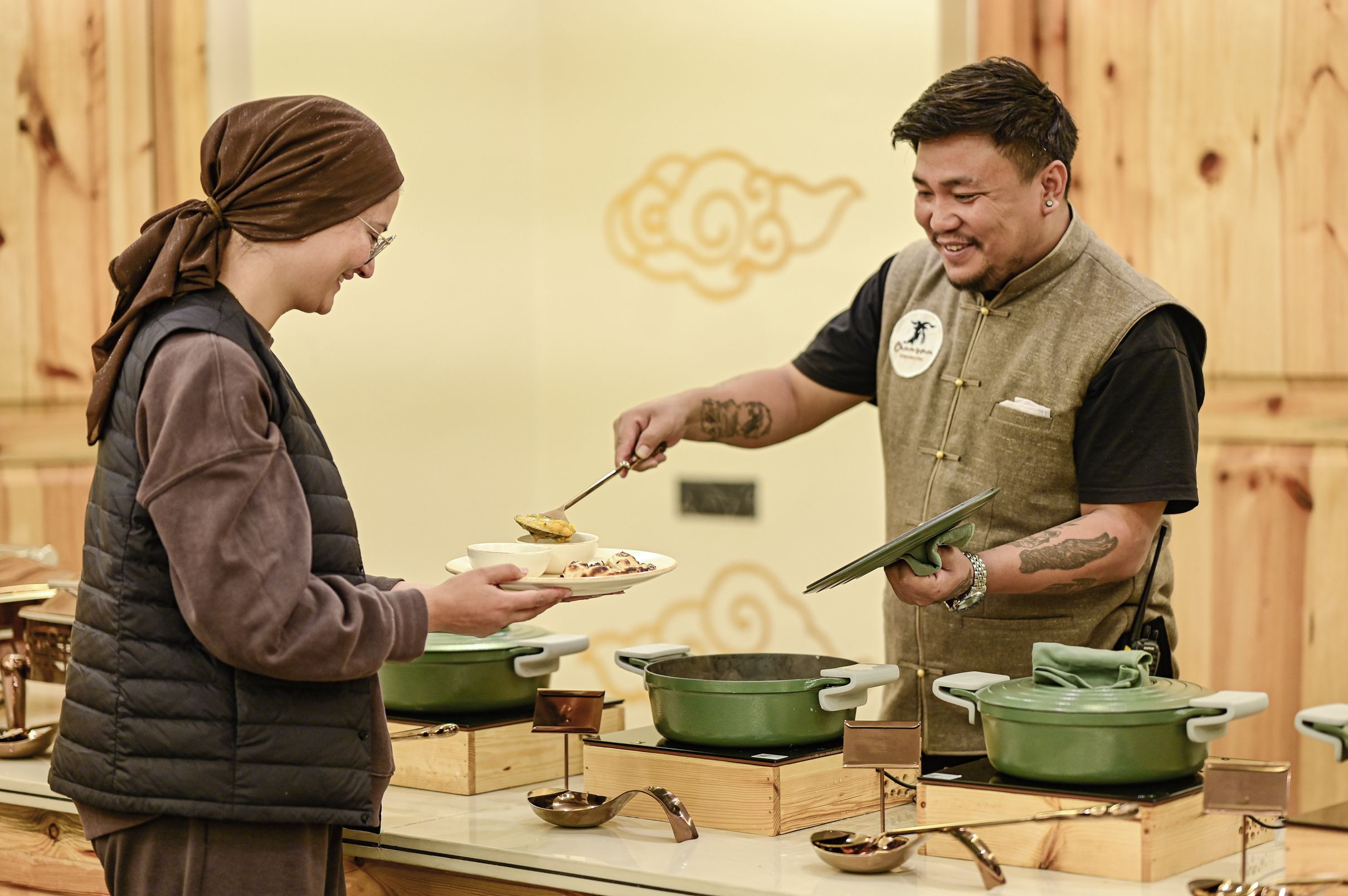 A person serving food to another person in a kitchen setting with wooden walls and various cooking utensils.