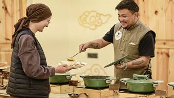 A person serving food to another person in a kitchen setting with wooden walls and various cooking utensils.