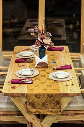 A wooden dining table set with plates, cutlery, and a vase with dried flowers. - Changpa Lodge (Leh)