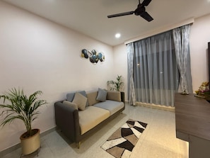 A living room with a sofa, a ceiling fan, a large window with curtains, and a decorative wall piece. - Atithi Guest House (Guwahati)