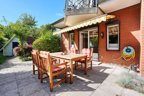 A patio with a wooden table and chairs, a brick building with a window, and a garden with trees and shrubs. - La Maison Eichenweg App 1 + 3 - Townhouse on Eichenweg (Scharbeutz)