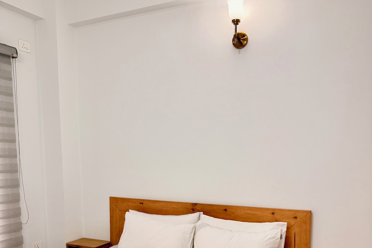 Una cama bien hecha con ropa blanca y una toalla doblada, cabecera de madera, mesitas de noche y una lámpara fijada a la pared.
