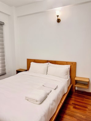 A neatly made bed with white linens and a folded towel, wooden headboard, bedside tables, and a wall-mounted light fixture. - Shetal Living Hattigauda (Burhanilkantha)