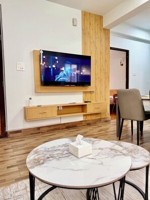 Habitación de hotel moderna con un televisor de pantalla plana montado en una pared de paneles de madera, una mesa con cubierta de mármol y una caja de pañuelos, y piso de madera.