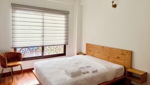 A bedroom with a bed, a chair, a nightstand, and a window with blinds. - Shetal Living Hattigauda (Burhanilkantha)