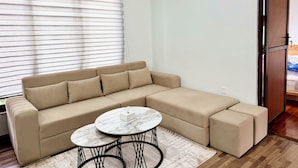 Een moderne woonkamer met een beige hoekbank, een ronde salontafel en een groot raam met zonwering.
