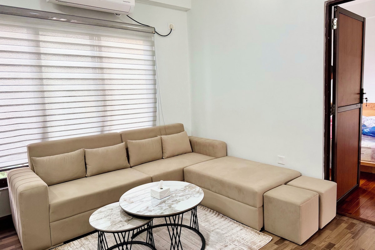 Una sala de estar moderna con un sofá seccional color beige, una mesa de centro redonda y un ventanal con persianas.