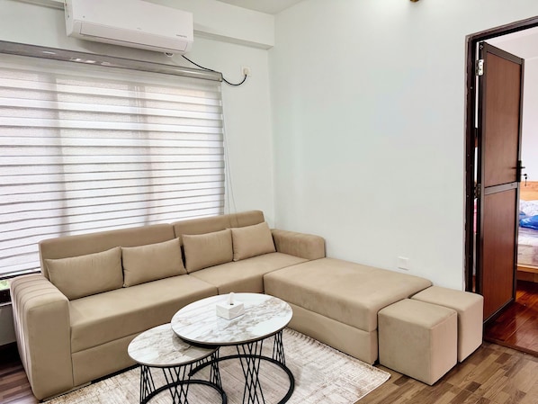 A modern living room with a beige sectional sofa, a round coffee table, and a large window with blinds. - Shetal Living Hattigauda (Burhanilkantha)