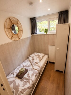A compact room with a single bed, a wardrobe, a window with curtains, and a wall-mounted mirror. - Zenda's natuurhuisje (Lanaken)