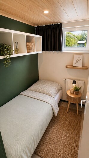 A compact room with a single bed, a small table with a lamp, a shelf with decorative items, and a window with a view. - Zenda's vakantiehuisje (Lanaken)