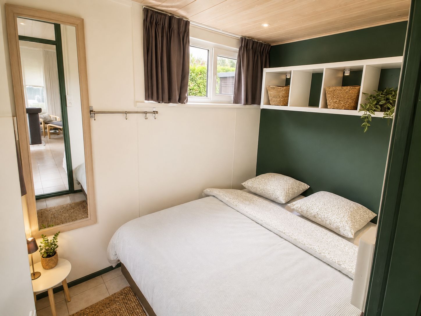 A bedroom with a bed, a mirror, a window with curtains, a bedside table with a lamp, and a shelf with baskets.