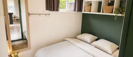 A bedroom with a bed, a mirror, a window with curtains, a bedside table with a lamp, and a shelf with baskets.
