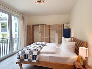 A bedroom with a bed, bedside table, lamp, and a wardrobe with a view to the outside. - Kogge 6 Apartment - Kogge Vacation Rental (Sassnitz)