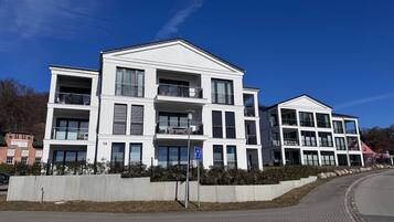 A modern multi-story residential building with balconies, a paved sidewalk, and a clear blue sky.