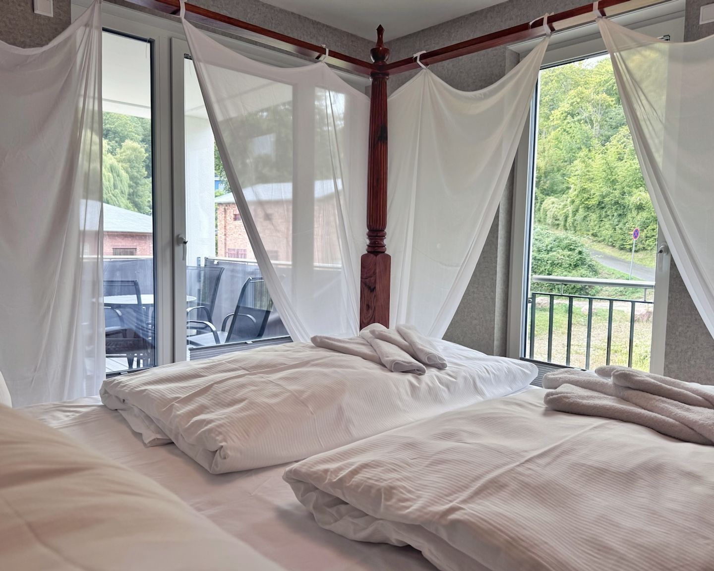 A bedroom with a four-poster bed, white bedding, and a view of a balcony and outdoor area.