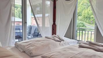 A bedroom with a four-poster bed, white bedding, and a view of a balcony and outdoor area.