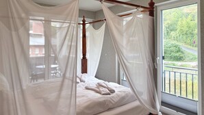 Uma cama com dossel e cortina, janela com vista para o verde e luminária no teto.