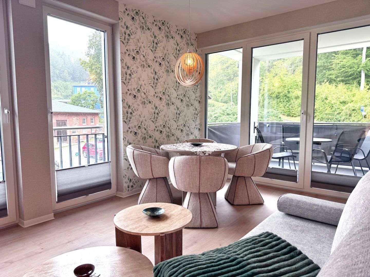 Ein modernes Wohnzimmer mit einem runden Tisch, zwei Stühlen, einer Couch und einem großen Fenster mit Blick ins Grüne.