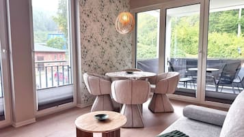 A modern living room with a round table, two chairs, a sofa, and a large window with a view of greenery.