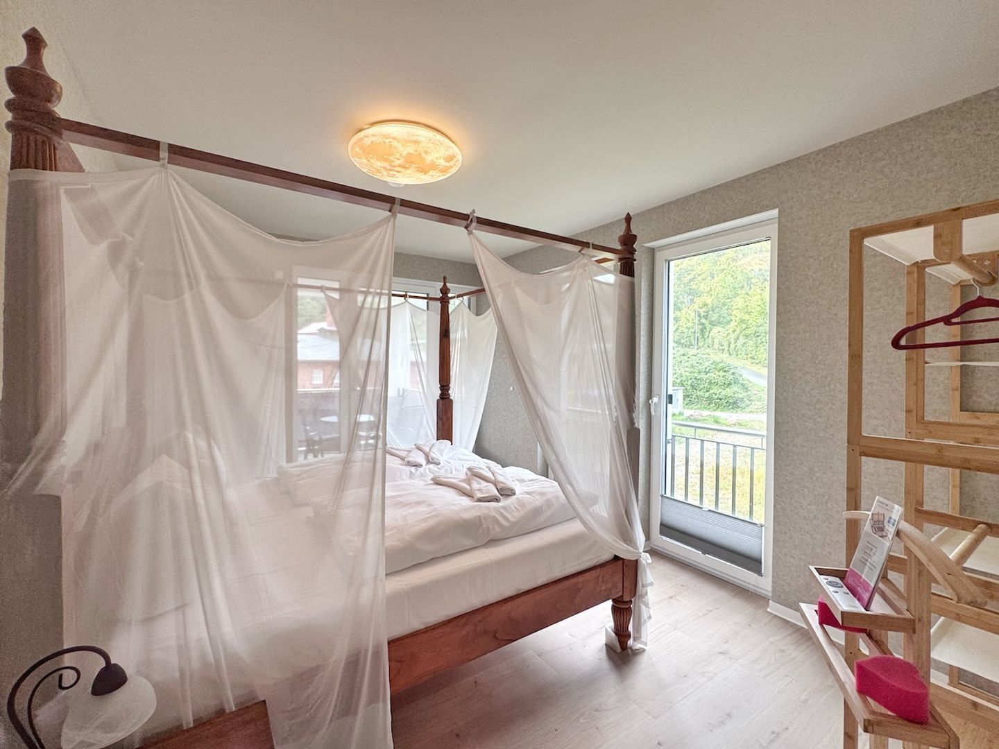A four-poster bed with a canopy, a wooden headboard, a bedside table with a red cushion, and a view of greenery outside.