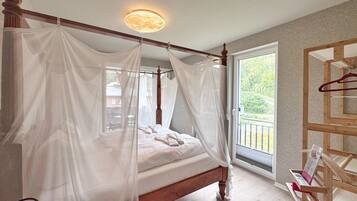 A four-poster bed with a canopy, a wooden headboard, a bedside table with a red cushion, and a view of greenery outside.