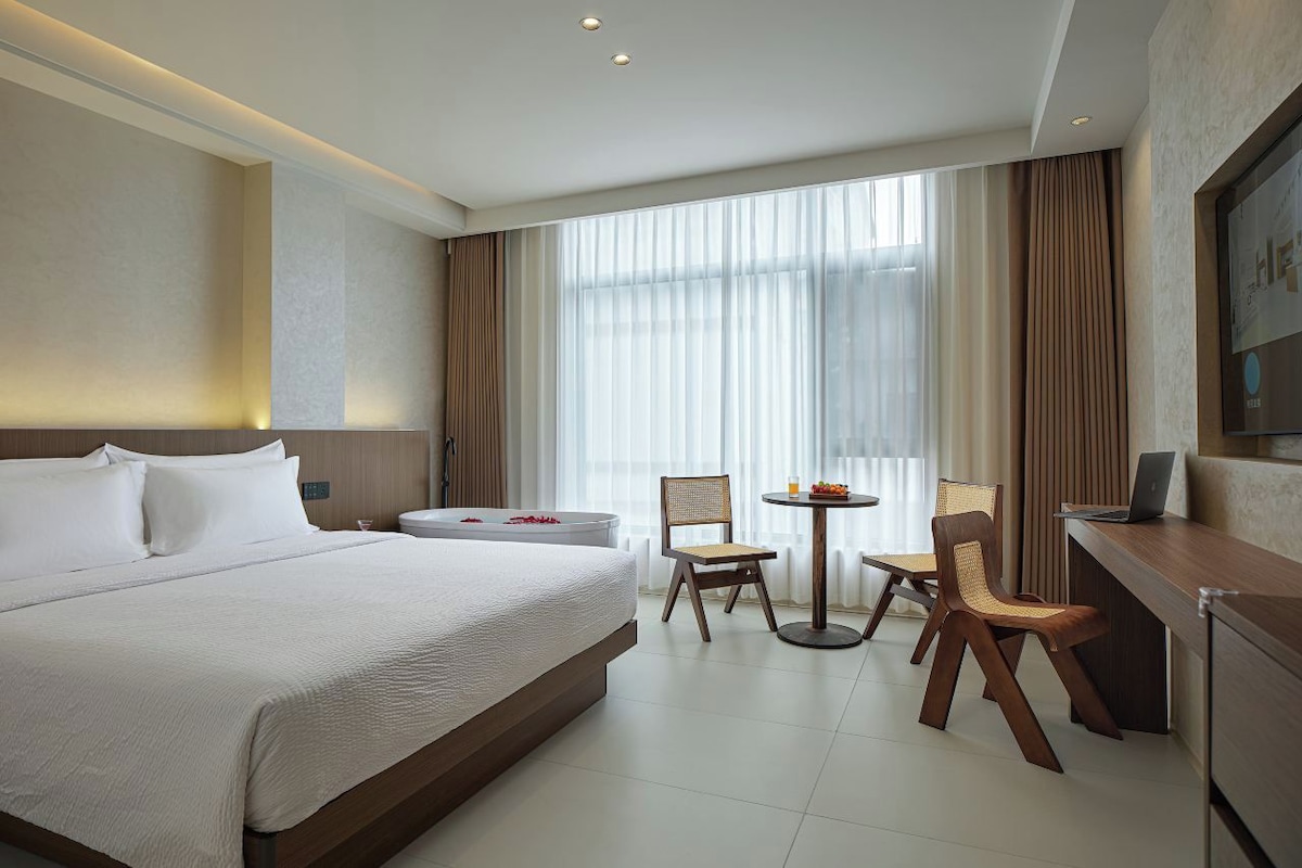 Habitación de hotel con una cama grande, un escritorio con una computadora portátil, una silla y una mesita con fruta.