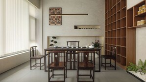Un coin repas avec une table et des chaises en bois, une étagère murale agrémentée d’objets décoratifs et une grande fenêtre avec des stores.