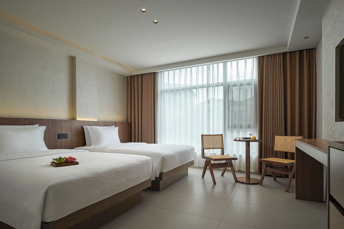 Habitación de hotel con dos camas, una mesita con una silla y un gran ventanal con cortinas.