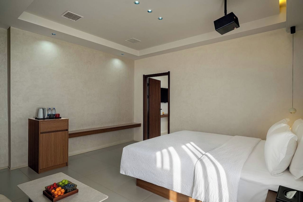 Una habitación de hotel moderna con una cama, un pequeño mueble de madera, un frutero y un proyector en el techo.
