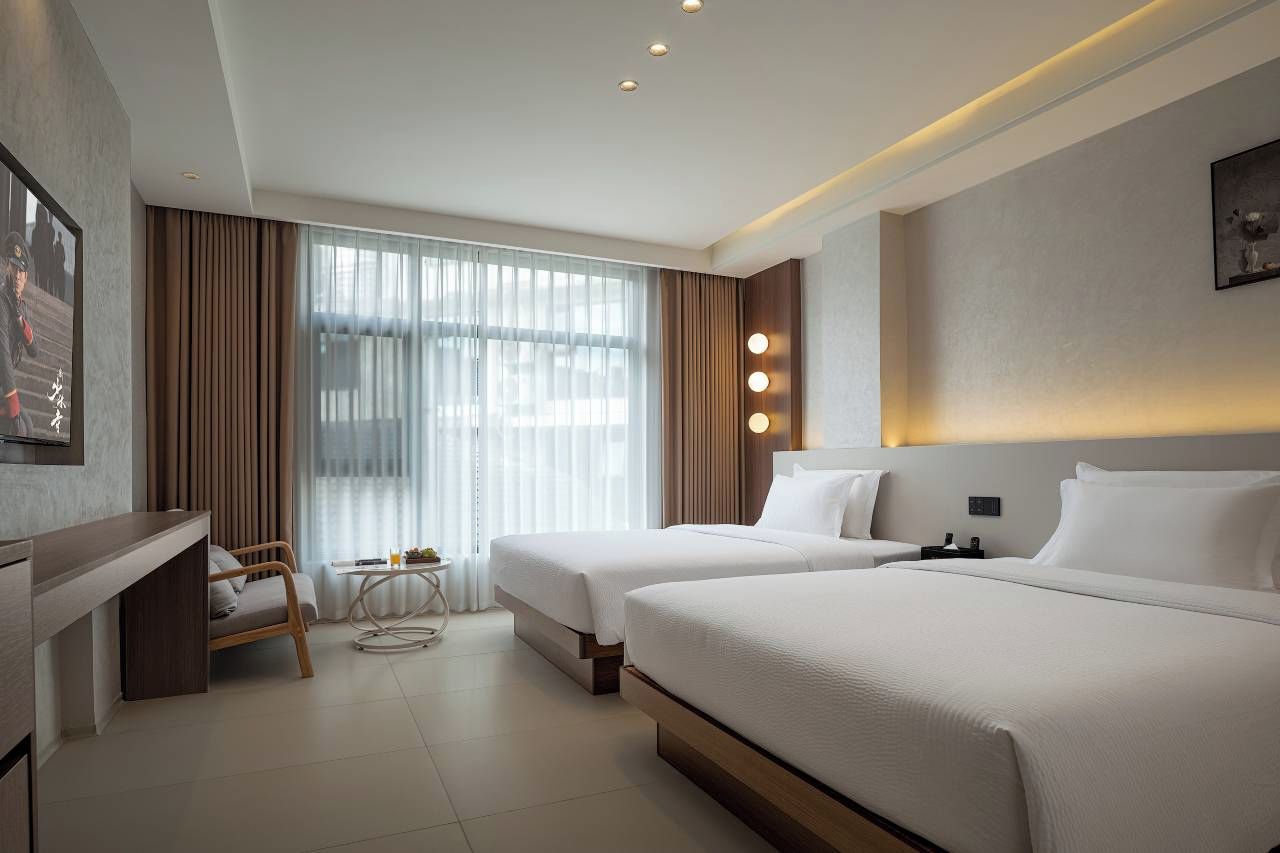 A hotel room with two beds, a desk, a chair, a small table, and a large window with curtains.