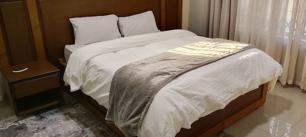 A neatly made bed with white linens and a grey throw blanket, a wooden nightstand with a drawer, and a window with curtains.