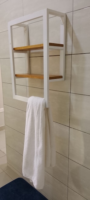 A bathroom with a white towel hanging on a rack, a mirror with a wooden shelf, and tiled walls.