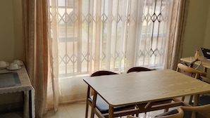 Ein Essbereich mit Tisch und Stühlen, eine Kaffeestation mit Tassen und Wasserkocher sowie ein Fenster mit dekorativen Vorhängen.