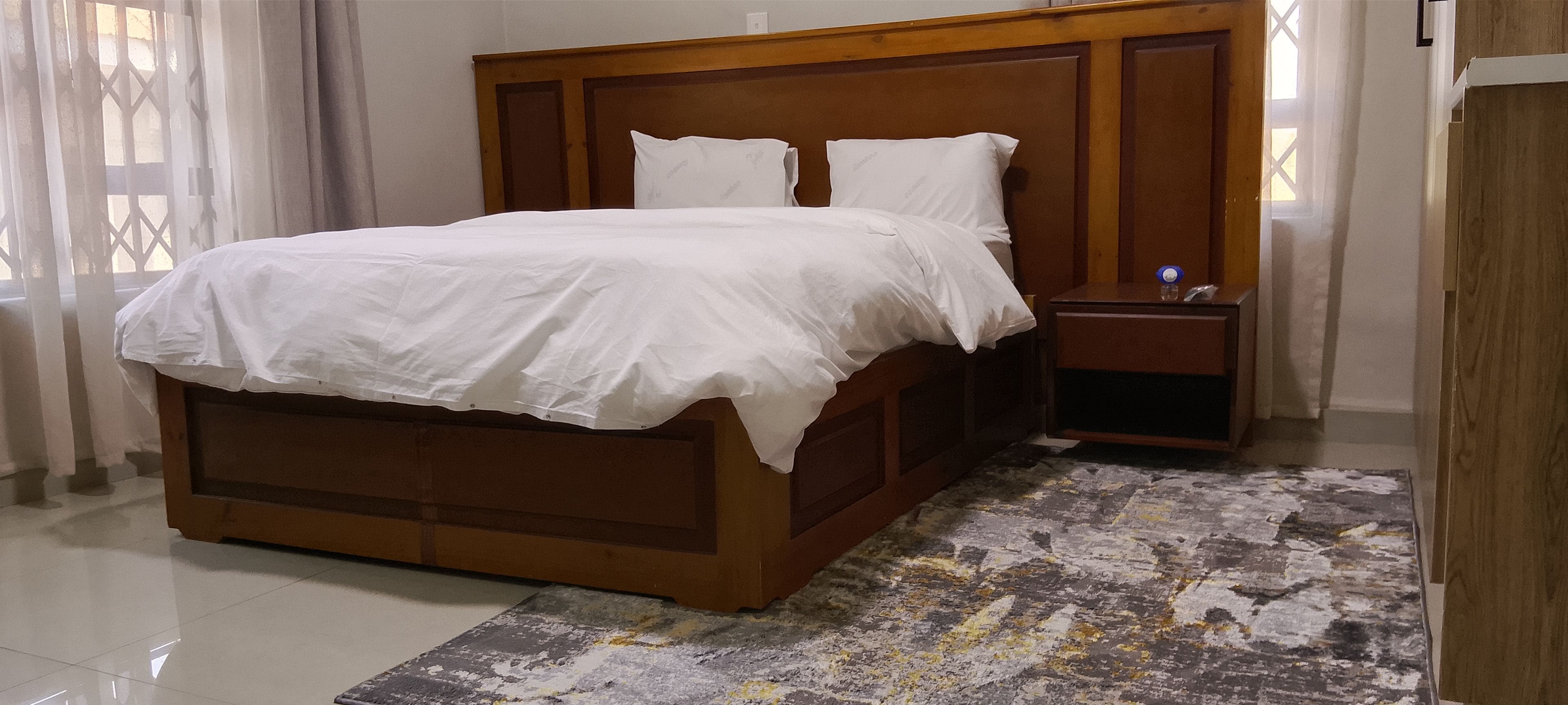 A bedroom with a wooden bed frame, white bedding, a nightstand, and a patterned rug.
