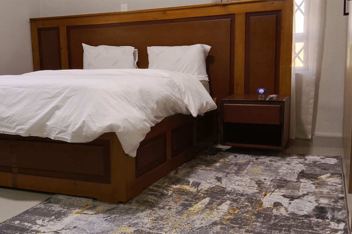 Un dormitorio con estructura de cama de madera, ropa de cama blanca, una mesita de noche y una alfombra estampada.